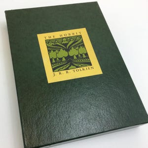 May include: A dark green book titled "The Hobbit" by J.R.R. Tolkien. The book has a textured cover and a yellow square label with the title and author's name. The label features a green illustration of trees and mountains.