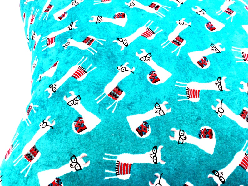 May include: A bright turquoise fabric featuring a repeating pattern of white llamas wearing black-rimmed glasses. Each llama is adorned with a small, colourful saddle blanket in shades of red, blue, and black, arranged in various orientations.