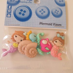 May include: A package of decorative buttons featuring mermaid and seashell designs. The buttons are in various colors, including pink, brown, and blue. The package includes the text "Mermaid Kisses" and the brand "Dress It Up".
