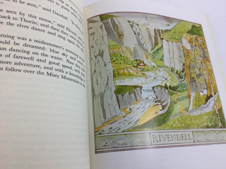 May include: A book page featuring a colorful illustration of a landscape with mountains, a river, and a small building. The word "RIVENDELL" is written at the bottom of the image. The opposite page contains printed text.