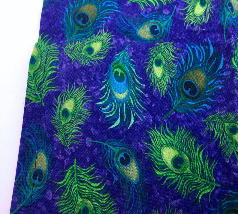 May include: Close-up of a fabric with a repeating peacock feather pattern. The feathers are in shades of green, blue, and gold against a deep purple background. The design is detailed and vibrant, suggesting a textile or decorative item.