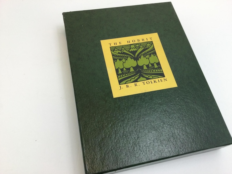 May include: A dark green book titled "The Hobbit" by J.R.R. Tolkien. The book has a textured cover and a yellow square label with the title and author's name. The label features a green illustration of trees and mountains.