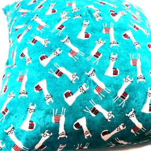May include: A square throw cushion featuring a vibrant turquoise background with a repeating pattern of white llamas. Each llama is depicted wearing black-rimmed glasses and a colourful patterned saddle blanket in shades of red, blue, and white.