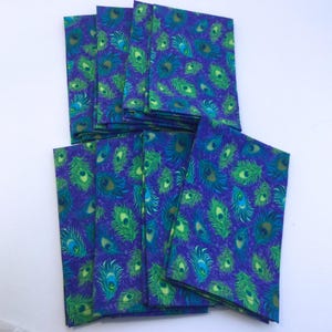 May include: A collection of folded fabric napkins with a vibrant peacock feather pattern. The napkins feature a deep blue background with green and turquoise feather designs. The napkins are stacked and arranged on a white surface.