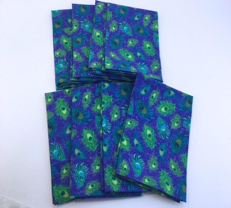 May include: A collection of folded fabric napkins with a vibrant peacock feather pattern. The napkins feature a deep blue background with green and turquoise feather designs. The napkins are stacked and arranged on a white surface.