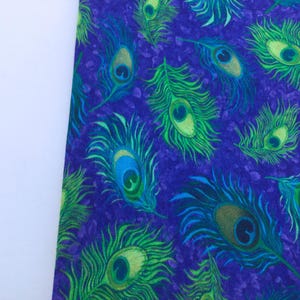 May include: Close-up of a fabric with a repeating peacock feather pattern. The feathers are in shades of green, blue, and yellow against a deep purple background. The fabric has a textured appearance.