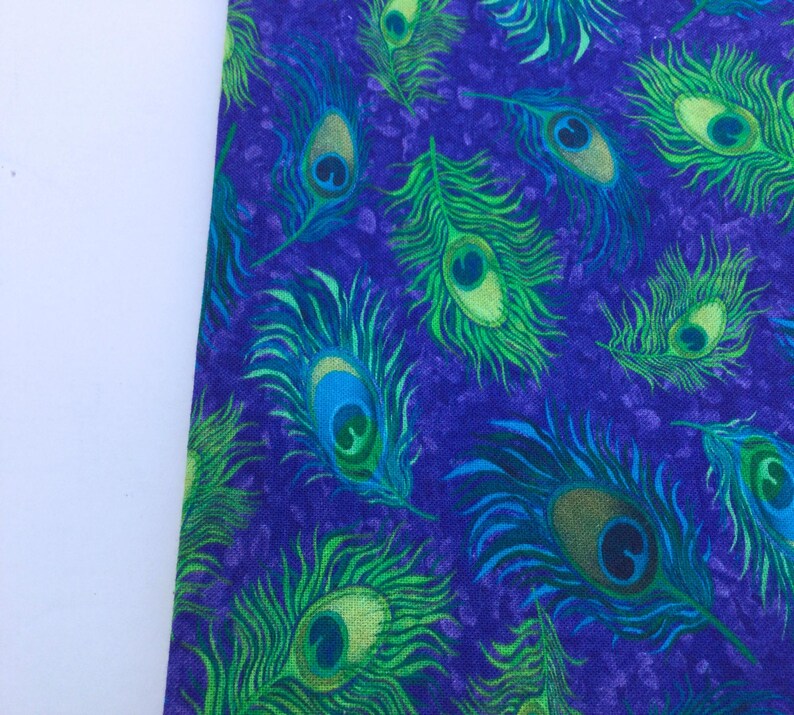 May include: Close-up of a fabric with a repeating peacock feather pattern. The feathers are in shades of green, blue, and yellow against a deep purple background. The fabric has a textured appearance.