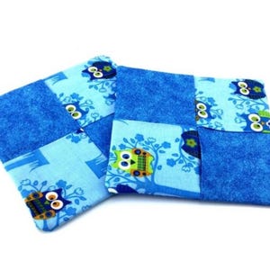 May include: Two square fabric coasters featuring a patchwork design. The pattern alternates between solid royal blue textured fabric and a light blue background printed with stylized owls in shades of green, blue, and yellow perched on trees.
