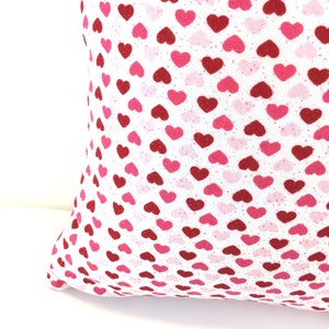 May include: A square throw pillow featuring a repeating pattern of small hearts in shades of red, dark pink, and light pink printed on a white fabric background. The textile has a visible woven texture and the hearts are arranged in a dense grid.