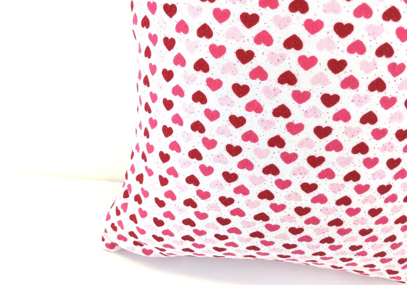 May include: A square throw pillow featuring a repeating pattern of small hearts in shades of red, dark pink, and light pink printed on a white fabric background. The textile has a visible woven texture and the hearts are arranged in a dense grid.