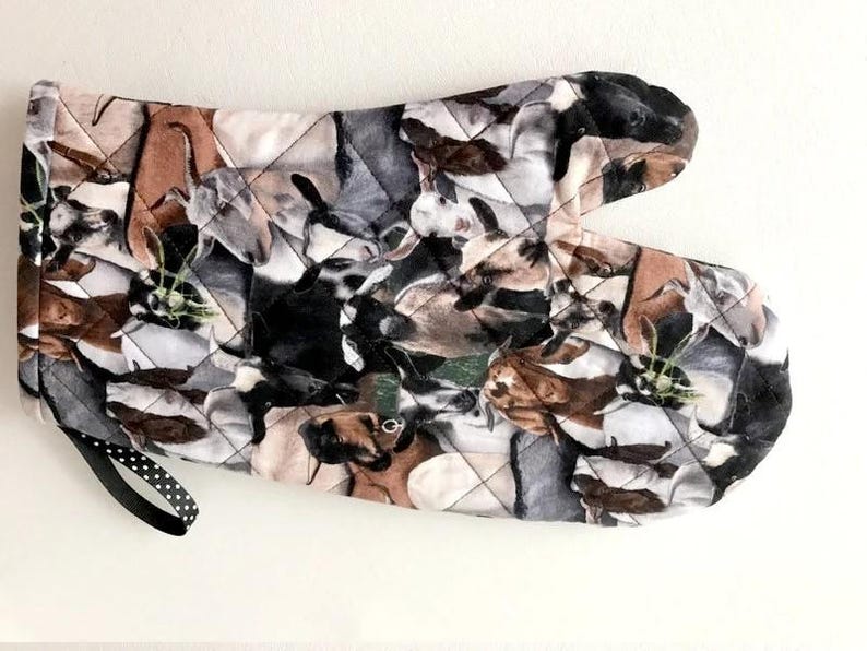 K&ouml;nnte beinhalten: Ein gesteppter Ofenhandschuh mit einem wiederkehrenden Ziegenmuster in verschiedenen Braun-, Schwarz-, Wei&szlig;- und Graut&ouml;nen. Der Stoff ist rautenf&ouml;rmig gesteppt und verf&uuml;gt &uuml;ber eine schwarze Schlaufe mit wei&szlig;en Punkten zum Aufh&auml;ngen an der Seite.