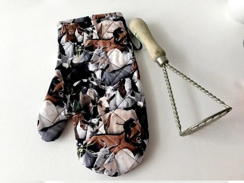 K&ouml;nnte beinhalten: Ein gesteppter Ofenhandschuh mit einem sich wiederholenden Muster aus braunen, schwarzen, wei&szlig;en und grauen Ziegen in verschiedenen Posen, kombiniert mit einem Kartoffelstampfer aus Metall mit hellem Holzgriff und gedrehtem Drahtrahmen.