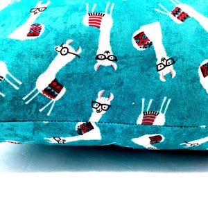 May include: A teal fabric cushion cover featuring a repeating pattern of white llamas wearing black-rimmed glasses and colourful patterned blankets. The llamas are oriented in various directions across the vibrant, textured teal background.