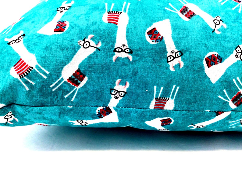 May include: A teal fabric cushion cover featuring a repeating pattern of white llamas wearing black-rimmed glasses and colourful patterned blankets. The llamas are oriented in various directions across the vibrant, textured teal background.