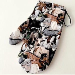 K&ouml;nnte beinhalten: Ein gesteppter Ofenhandschuh mit einem sich wiederholenden Muster aus Ziegen in verschiedenen Farben wie Braun, Schwarz, Wei&szlig; und Grau. Der Stoff hat eine rautenf&ouml;rmige Steppung und eine schwarze Stoffschlaufe zum Aufh&auml;ngen des Handschuhs.