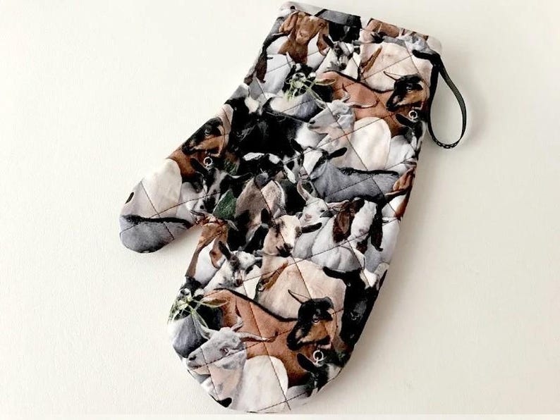 K&ouml;nnte beinhalten: Ein gesteppter Ofenhandschuh mit einem sich wiederholenden Muster aus Ziegen in verschiedenen Farben wie Braun, Schwarz, Wei&szlig; und Grau. Der Stoff hat eine rautenf&ouml;rmige Steppung und eine schwarze Stoffschlaufe zum Aufh&auml;ngen des Handschuhs.