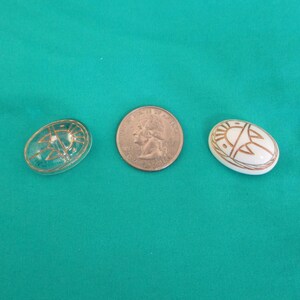 May include: Two decorative oval objects and a coin are displayed. One object is clear with a gold design, the other is white with a gold design. The objects are on a teal background.