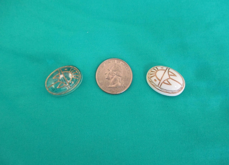 May include: Two decorative oval objects and a coin are displayed. One object is clear with a gold design, the other is white with a gold design. The objects are on a teal background.