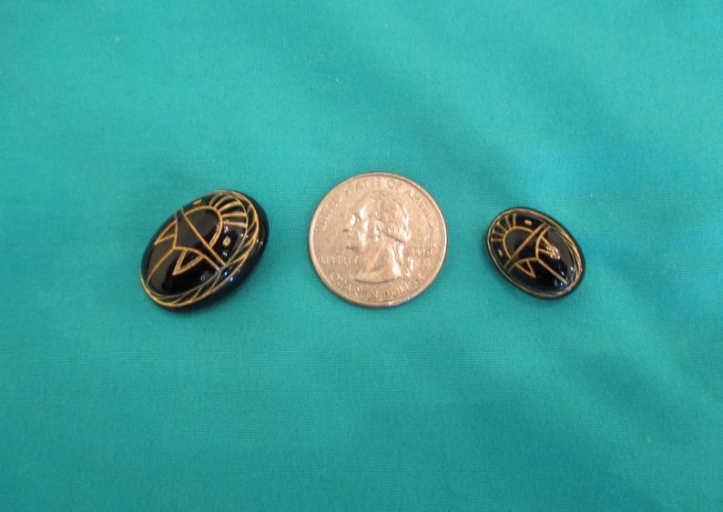 May include: Two black oval buttons with gold designs, and a U.S. quarter coin. The buttons feature a sun-like design. The buttons are on teal fabric.