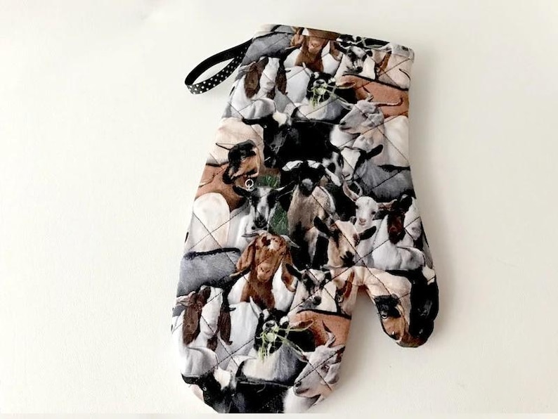 K&ouml;nnte beinhalten: Ein gesteppter Ofenhandschuh mit einem sich wiederholenden Muster aus verschiedenen Ziegen in Braun-, Schwarz-, Wei&szlig;- und Graut&ouml;nen. Der Handschuh verf&uuml;gt &uuml;ber eine schwarze Stoffschlaufe mit kleinen wei&szlig;en Punkten zur praktischen Aufbewahrung.