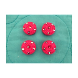 Puede incluir: Cuatro botones circulares de color rosa brillante dispuestos en formación cuadrada. Cada botón presenta un patrón geométrico pentagonal en relieve en su superficie, acentuado con pequeños cristales reflectantes multicolores incrustados.