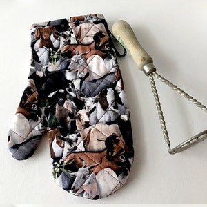 May include: A quilted oven mitt with a pattern of goats and sheep. The mitt is brown, black, and white. A silver potato masher is also in the image.