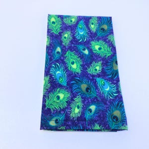 May include: A folded fabric item with a vibrant peacock feather pattern. The design features green, blue, and gold feather details on a deep purple background. The fabric is likely cotton or a similar material, suitable for various craft projects.