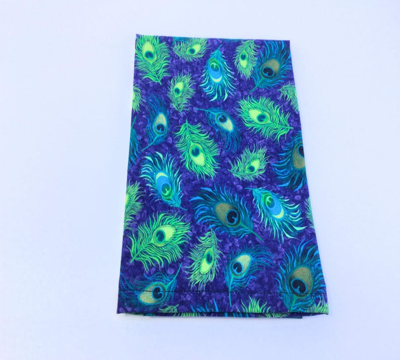 May include: A folded fabric item with a vibrant peacock feather pattern. The design features green, blue, and gold feather details on a deep purple background. The fabric is likely cotton or a similar material, suitable for various craft projects.