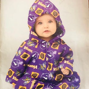 May include: A baby wearing a purple hooded jumpsuit with a zipper closure. The jumpsuit is patterned with football helmets, footballs, and the word "Vikings" in yellow and white. The pattern is on a purple background. The image is a Simplicity sewing pattern.