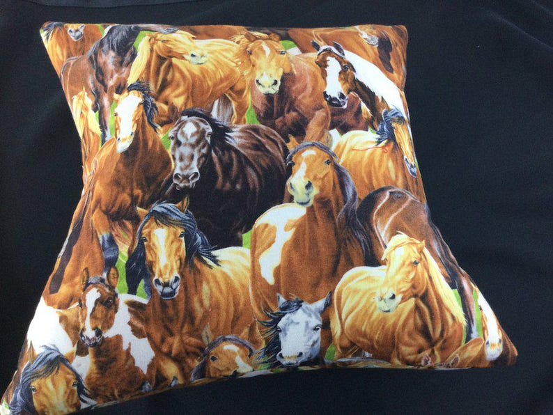 May include: A square throw pillow featuring a dense, colorful print of multiple horses in various shades of brown, tan, white, and black. The horses are depicted in close proximity, showing their heads and manes against a subtle green background.