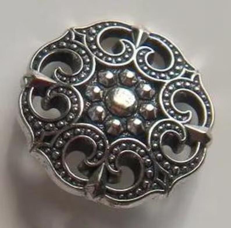 以下が含まれることがあります： 装飾的な装飾が施された銀色の金属ボタン。ボタンの中央には、テクスチャ加工された表面を持つ、隆起した円形の領域があります。