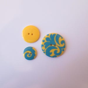 May include: Three yellow and blue decorative buttons. One solid yellow button with two holes, a small button with a blue swirl design, and a larger button with a blue floral pattern.