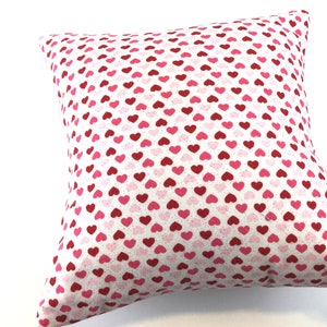 May include: A square throw pillow featuring a repeating pattern of small hearts in shades of red and pink against a white background. The fabric has a subtle speckled texture throughout, and the pillow is filled to create a plump, soft shape.