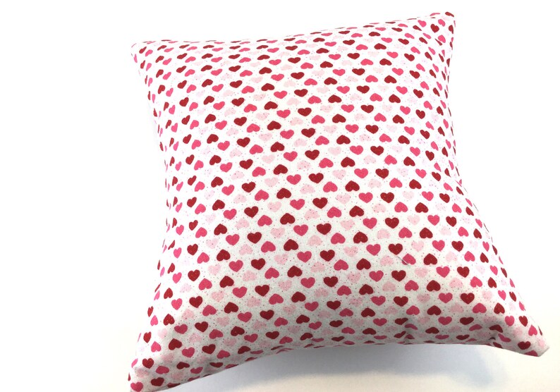 May include: A square throw pillow featuring a repeating pattern of small hearts in shades of red and pink against a white background. The fabric has a subtle speckled texture throughout, and the pillow is filled to create a plump, soft shape.