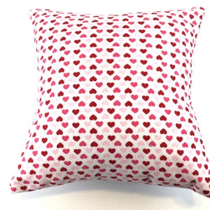 May include: A square throw pillow featuring a repeating pattern of small hearts in shades of red, dark pink, and light pink against a white background. The fabric has a subtle speckled texture throughout the design, covering the entire surface.