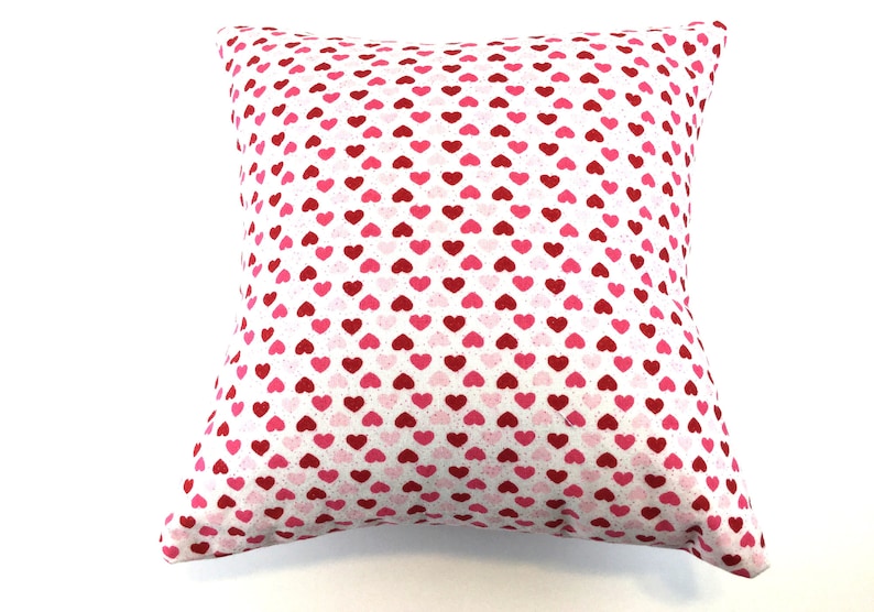 May include: A square throw pillow featuring a repeating pattern of small hearts in shades of red, dark pink, and light pink against a white background. The fabric has a subtle speckled texture throughout the design, covering the entire surface.