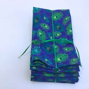 May include: A stack of folded fabric napkins with a peacock feather pattern in shades of blue, green, and yellow. The napkins are tied together with a green ribbon. The fabric has a textured appearance.