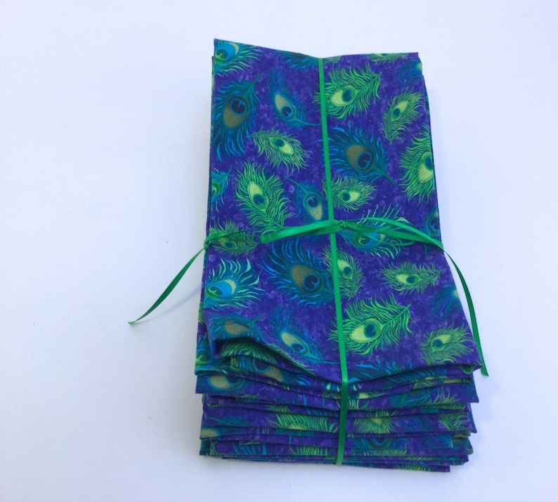 May include: A stack of folded fabric napkins with a peacock feather pattern in shades of blue, green, and yellow. The napkins are tied together with a green ribbon. The fabric has a textured appearance.