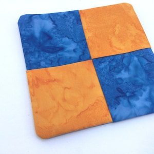 May include: A square fabric coaster with a patchwork design. The coaster is made of four squares of fabric, two blue and two orange. The fabric has a tie-dye pattern.