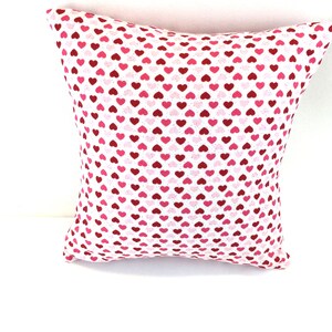 May include: A square throw pillow featuring a repeating pattern of small hearts in various shades of red, pink, and white. The fabric has a white base color, and the heart shapes are distributed evenly across the entire surface of the pillow.