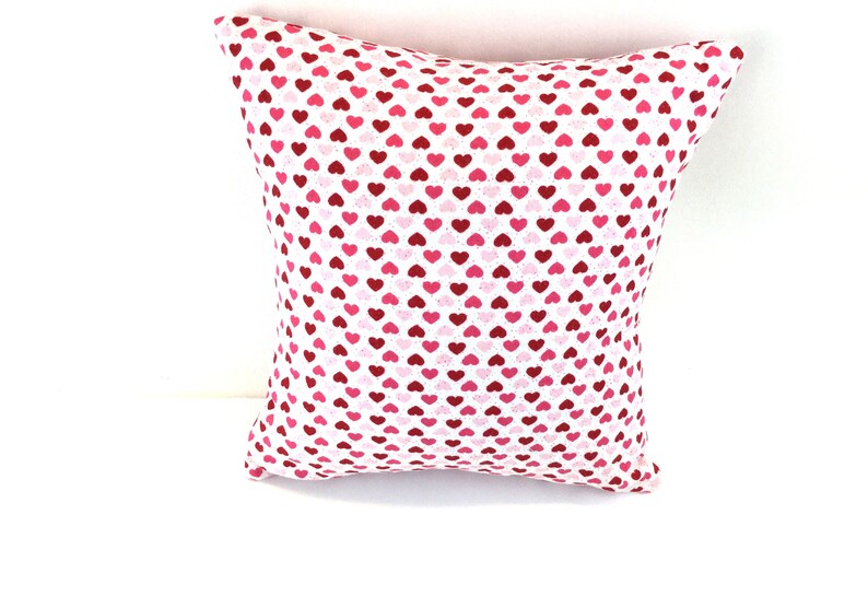 May include: A square throw pillow featuring a repeating pattern of small hearts in various shades of red, pink, and white. The fabric has a white base color, and the heart shapes are distributed evenly across the entire surface of the pillow.
