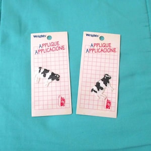 May include: Two Wrights brand applique patches featuring a black and white cow design. Each patch is mounted on a white rectangular card with a red grid pattern and the text APPLIQUE APPLICACIONE printed at the top in red and blue lettering.