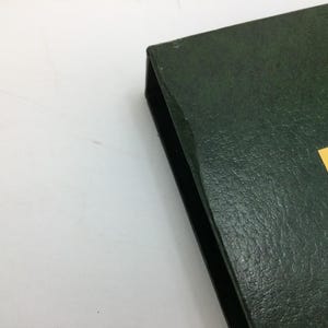 May include: A dark green, textured book or binder with a black spine. A yellow label with green and brown text is visible on the corner. The item is on a white surface.