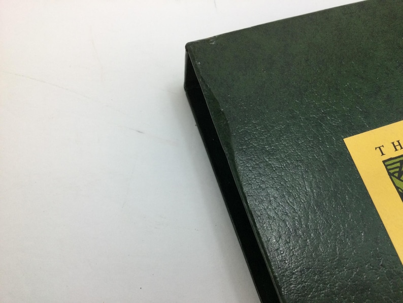 May include: A dark green, textured book or binder with a black spine. A yellow label with green and brown text is visible on the corner. The item is on a white surface.