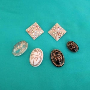 May include: A collection of decorative jewellery items, including two square, rhinestone-studded earrings, a clear oval, and three oval pendants with detailed designs. The pieces are on a teal background.