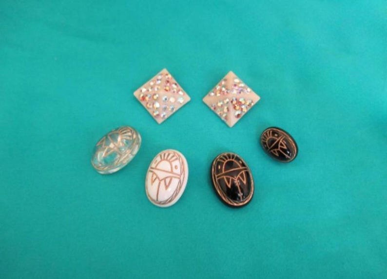 May include: A collection of decorative jewellery items, including two square, rhinestone-studded earrings, a clear oval, and three oval pendants with detailed designs. The pieces are on a teal background.