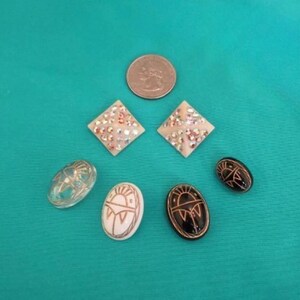 May include: A collection of jewellery items, including two diamond-shaped earrings with iridescent details, three oval earrings with sun designs, and a clear oval earring. A coin is also present.