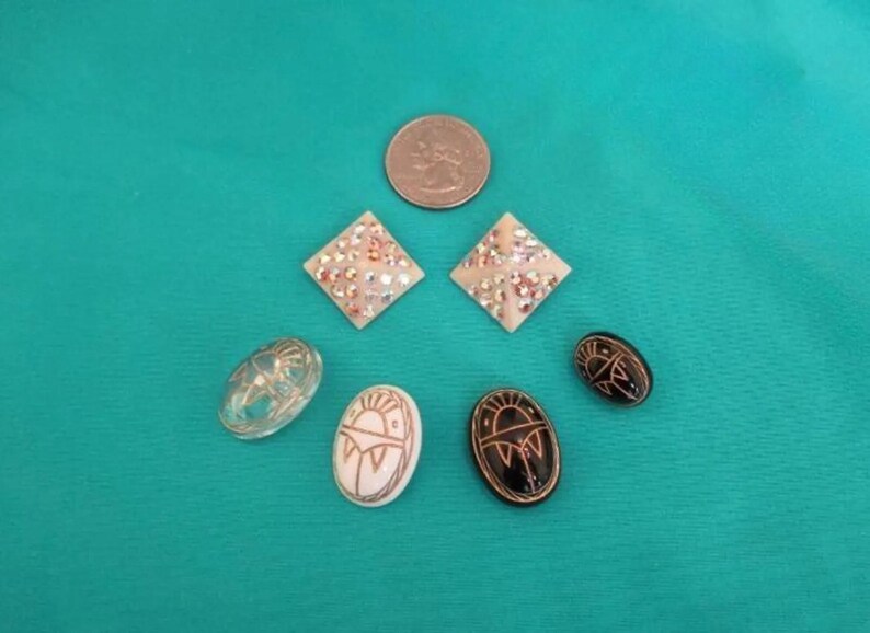 May include: A collection of jewellery items, including two diamond-shaped earrings with iridescent details, three oval earrings with sun designs, and a clear oval earring. A coin is also present.