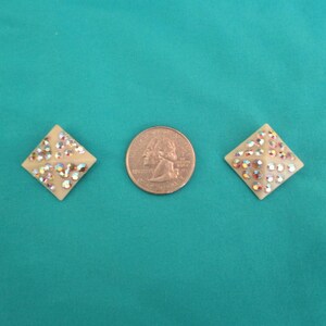 May include: Two cream-coloured, diamond-shaped earrings with iridescent rhinestones are displayed next to a quarter coin. The earrings are set against a teal background, showcasing their sparkling details.