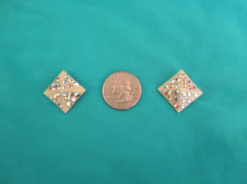 May include: Two cream-coloured, diamond-shaped earrings with iridescent rhinestones are displayed next to a quarter coin. The earrings are set against a teal background, showcasing their sparkling details.